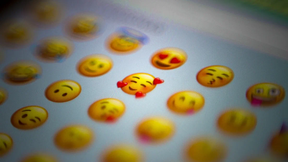 Close-up of a smartphone emoji keyboard, with a smiling face surrounded by three red hearts in focus among other facial-expression emojis.
