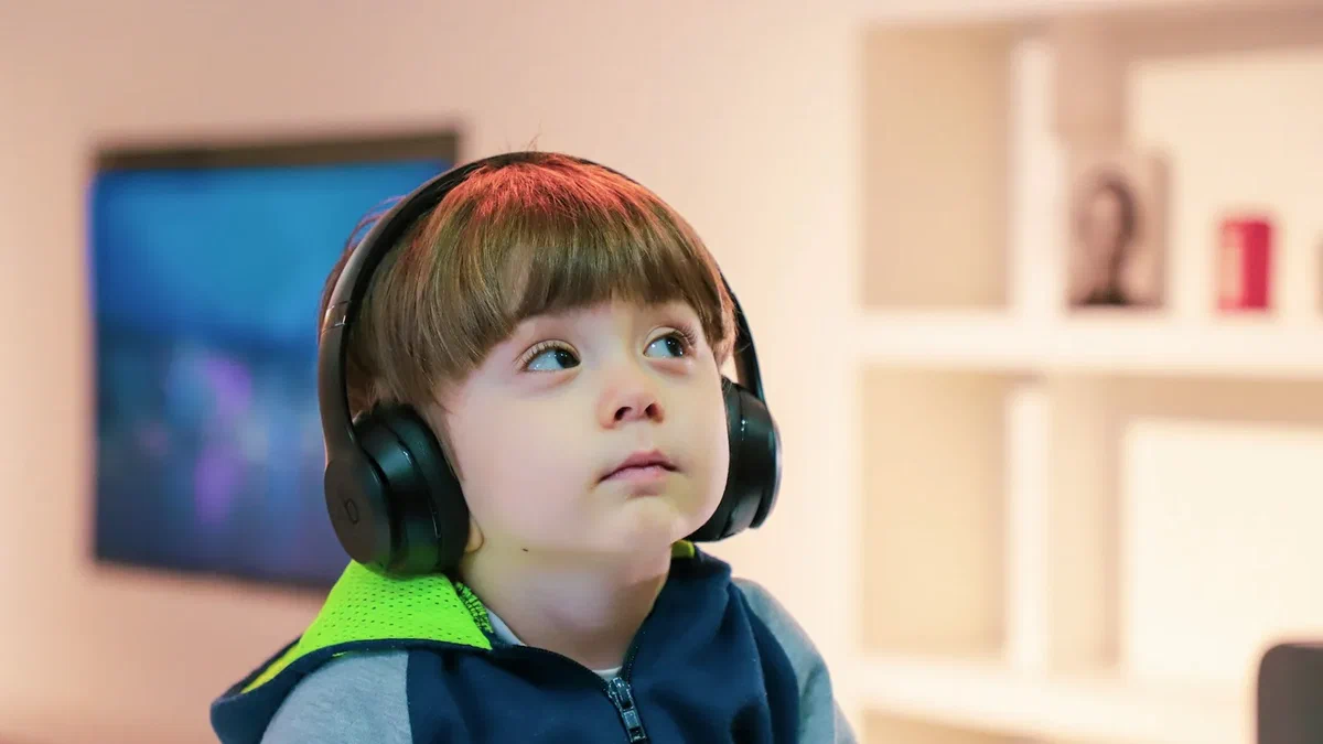 A young child wearing large black over-ear headphones, looking up and to one side, in a softly lit room with a blurred television screen behind them.