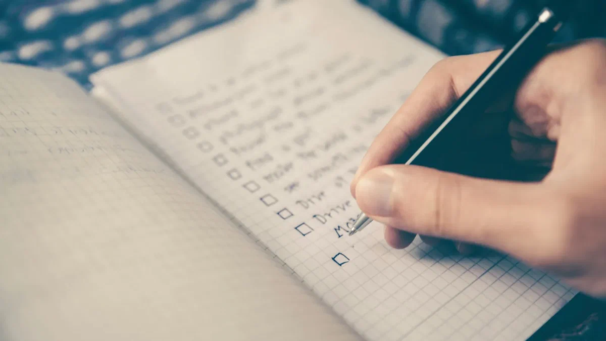 A close-up of a right hand holding a black pen mid-write, working down a handwritten to-do list of empty square tick boxes on the cross-ruled page of an open notebook resting on a dark patterned cloth, with a couple of the boxes further down the list already filled.