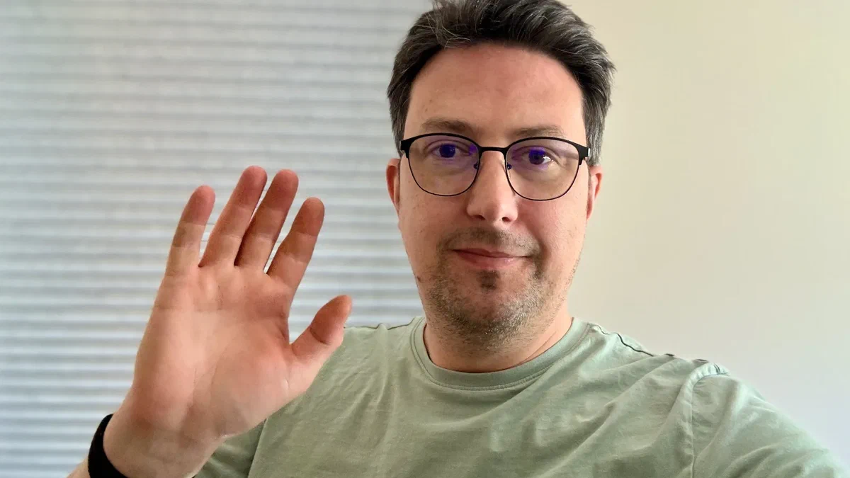 Axel, who has short dark hair, glasses and a trimmed beard, wearing a sage-green t-shirt and smiling gently while raising one hand in a wave, in a softly lit room with a window blind behind him.